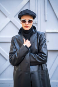 Person wearing a black leather coat, black turtleneck, white sunglasses, and a black cap, posing against a gray urban wall.