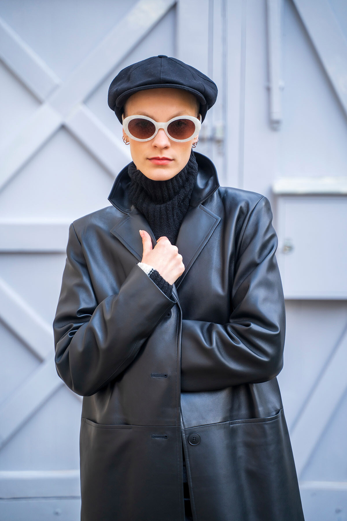 Person wearing a black leather coat, black turtleneck, white sunglasses, and a black cap, posing against a gray urban wall.