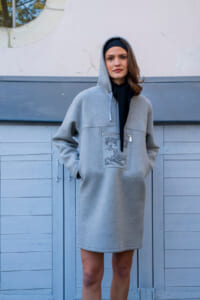 Woman in a gray hooded sweatshirt dress with hands in pockets, standing in front of blue wooden panels.