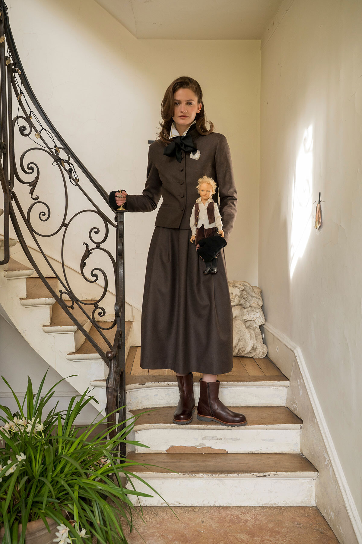 Woman in a vintage brown suit and black bow stands on a staircase, holding a small doll dressed in brown and white beside her.