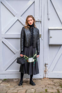 Woman in a black leather jacket and long printed skirt stands against a gray wooden door, holding a small black handbag.