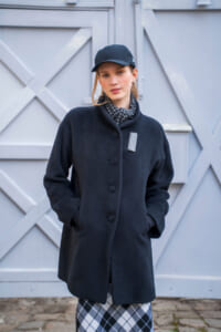 Woman in a navy wool coat with hands in pockets, wearing a black cap and patterned scarf, standing in front of a light gray wooden gate.