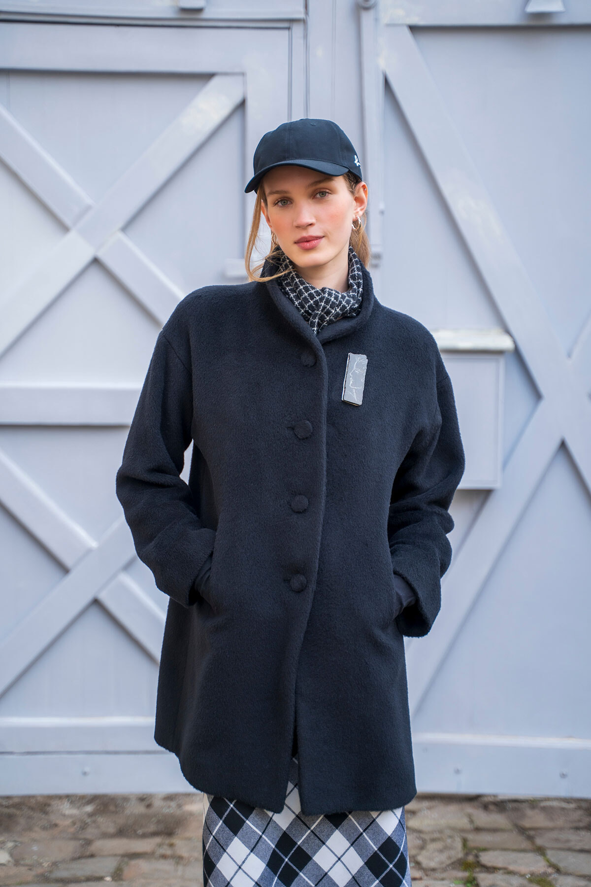 Woman in a navy wool coat with hands in pockets, wearing a black cap and patterned scarf, standing in front of a light gray wooden gate.