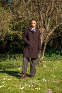 Person standing in a sunlit garden wearing a dark brown knee-length coat, gray scarf, gloves, gray trousers, and brown boots with a flowering tree behind.