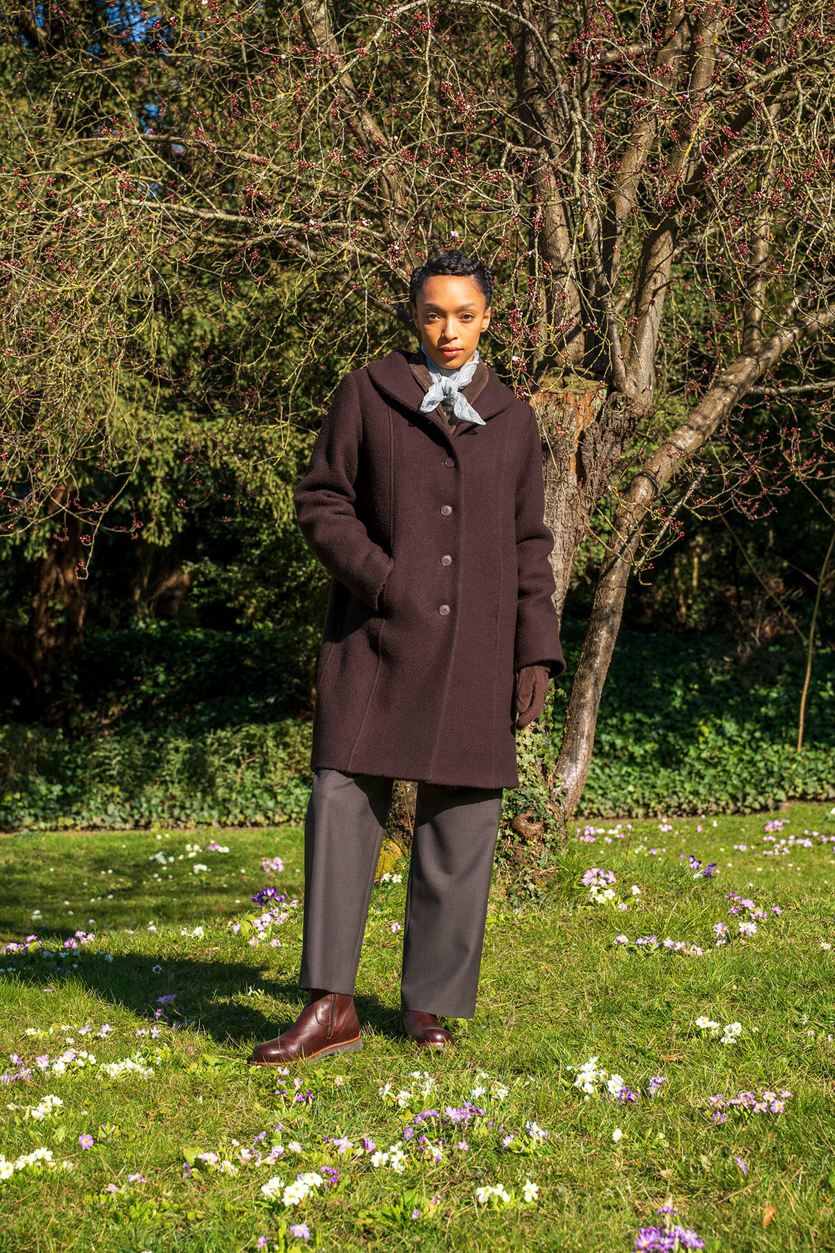 Person standing in a sunlit garden wearing a dark brown knee-length coat, gray scarf, gloves, gray trousers, and brown boots with a flowering tree behind.