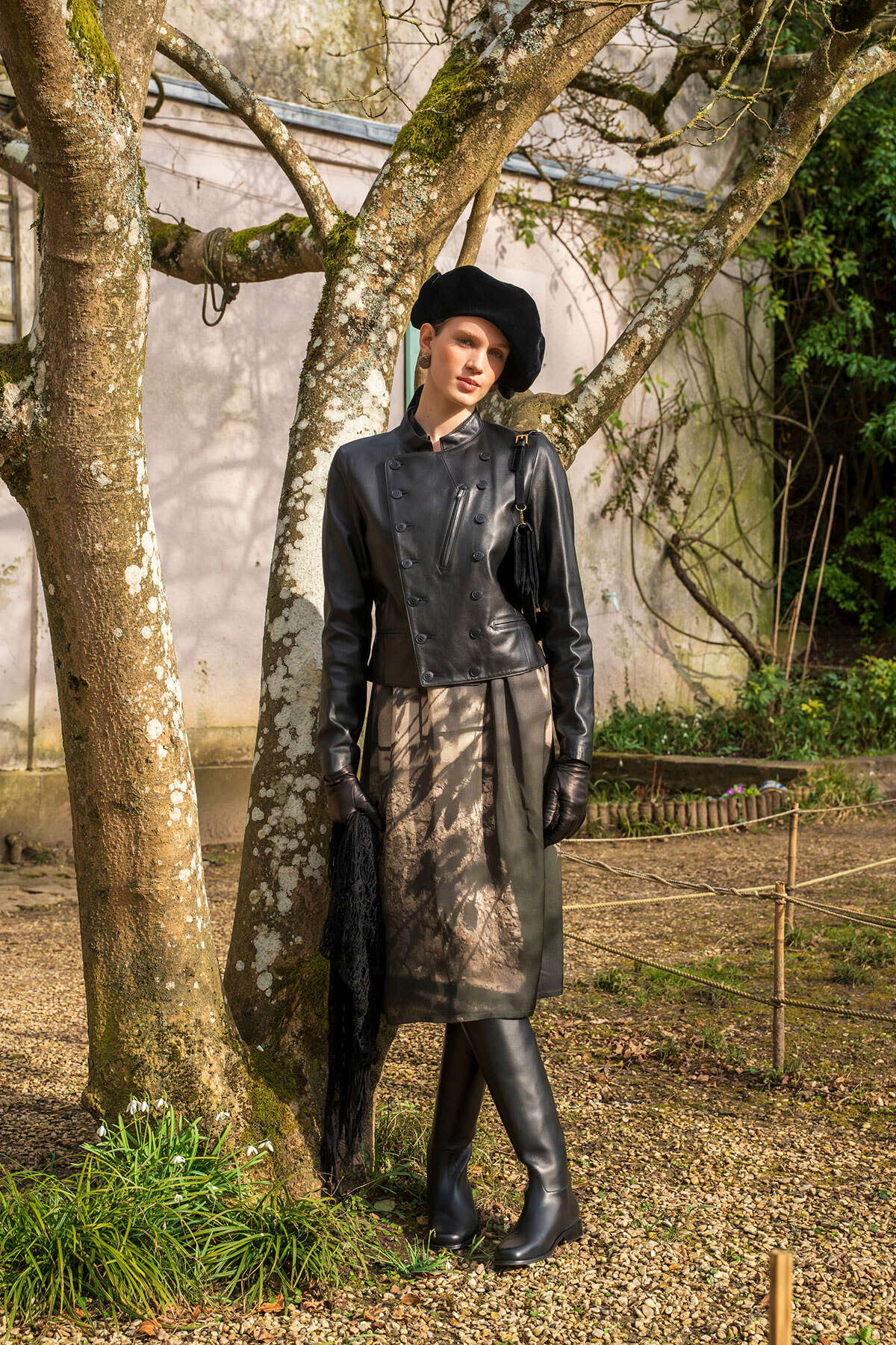 Woman in a black beret and double-breasted leather coat standing beside a tree in a sunlit garden, wearing gloves and knee-high boots with a black fringed bag over the shoulder.