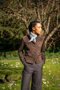 Woman standing in a sunlit garden wearing a brown suede blazer, gray scarf, and dark trousers, with blossoming branches in the background.