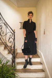 Woman in a black button-front midi dress with a waist tie and gloves, standing on a wooden staircase with a black handbag.