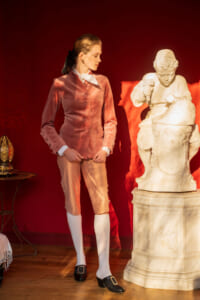 Person in a salmon-pink velvet 18th-century-style coat and breeches, white stock collar, white stockings, and black buckle shoes stands beside a white marble statue of a child sculpting a rock, against a red backdrop.