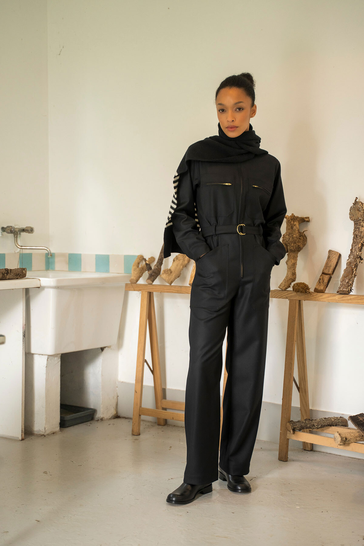 Woman in a black utility jumpsuit and scarf stands in a studio beside a workbench with carved logs on display tables.