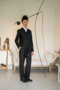 Woman in a black suit standing with hands in pockets in a minimalist room with a modern floor lamp and a bed in the background.