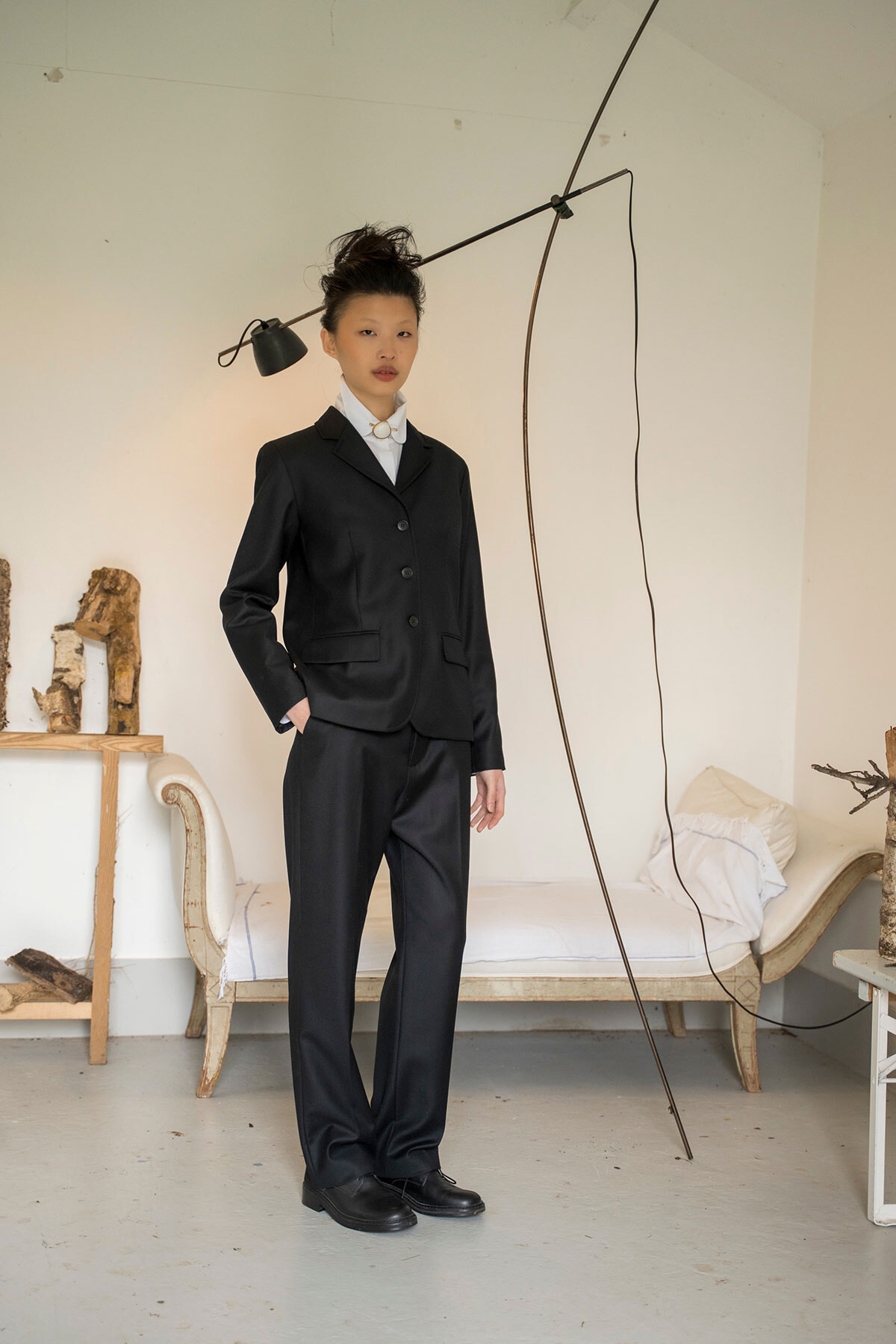 Woman in a black suit standing with hands in pockets in a minimalist room with a modern floor lamp and a bed in the background.