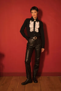 Person standing against a red wall in a black blouse with white ruffled front, black leather pants, and a large belt buckle.