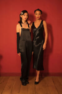 Two women pose against a red wall in black evening outfits: one in satin overalls with long black gloves and a pearl necklace, the other in a black slip dress with heels and a bracelet.