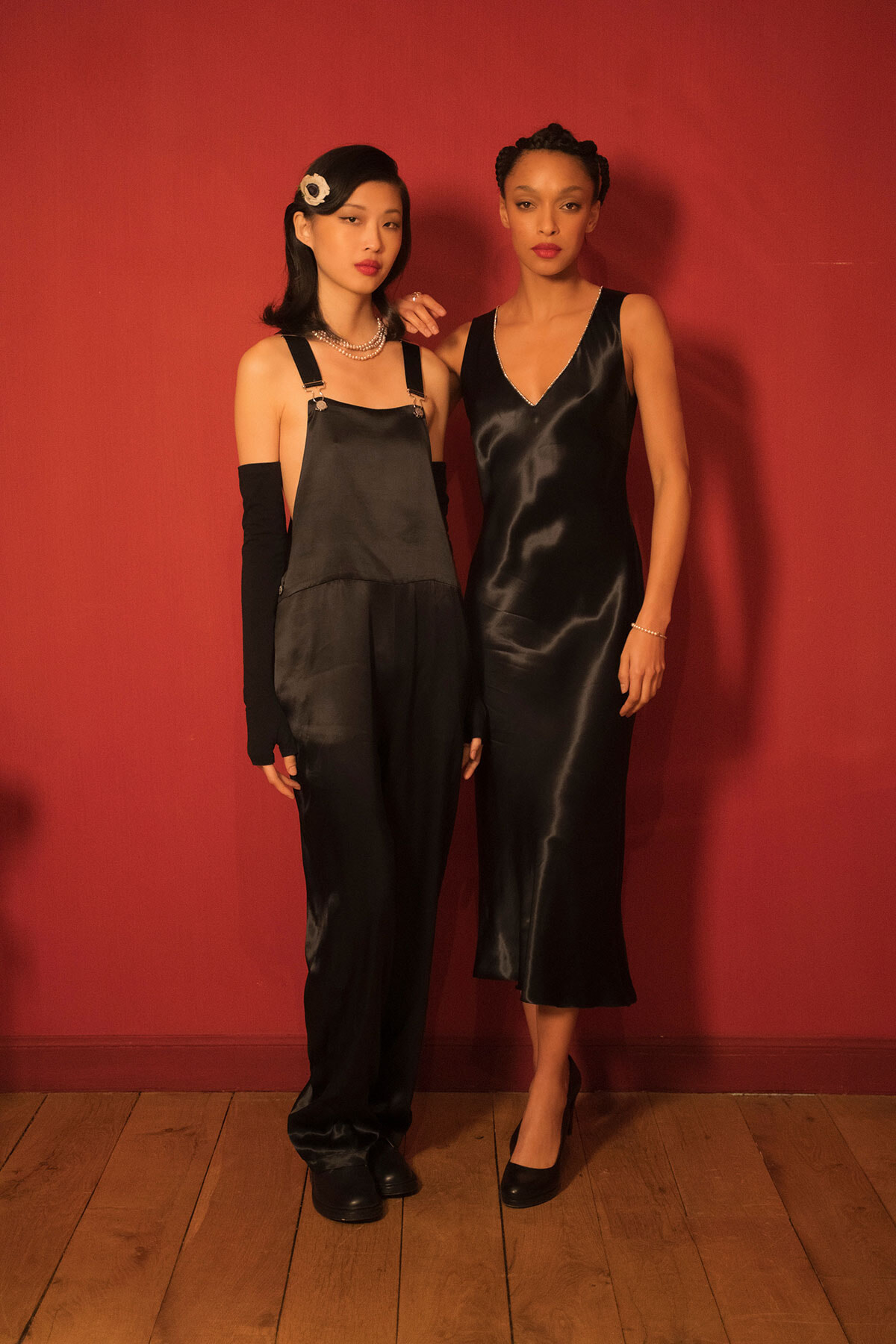 Two women pose against a red wall in black evening outfits: one in satin overalls with long black gloves and a pearl necklace, the other in a black slip dress with heels and a bracelet.