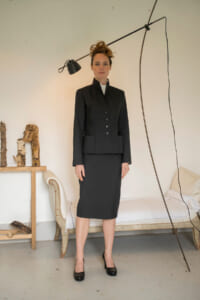 Woman in a tailored black blazer and knee-length skirt, wearing black heels, standing in a neutral room with a hanging lamp above.