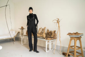 Person in black overalls and a beanie stands in an art studio, surrounded by carved wood pieces and driftwood on tables and benches.
