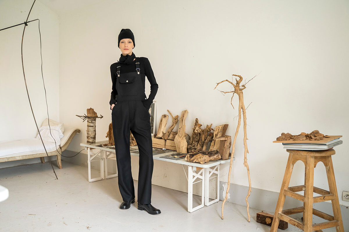 Person in black overalls and a beanie stands in an art studio, surrounded by carved wood pieces and driftwood on tables and benches.