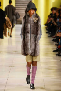 Runway model in a long gray fur vest with a gold brooch, dark hat, beige shorts, pink leg warmers, and black shoes.