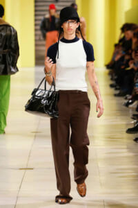 Model on a runway wearing a white sleeveless top with navy sleeves, brown trousers, brown loafers, a shiny black handbag, and a black hat with glasses.