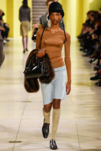Model walking the runway in a camel knit top, pale blue wrap skirt, beige knee-high socks, black hat, and glossy black bag with fur stole accessory.