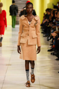 Model walking a fashion show runway in a peach textured blazer and matching skirt, with large lapels, a brooch, and patterned socks with brown shoes.