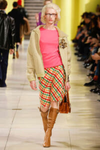 Female model on a fashion runway wearing a pink top, beige jacket, plaid shorts, tan knee-high boots, and a brown handbag.
