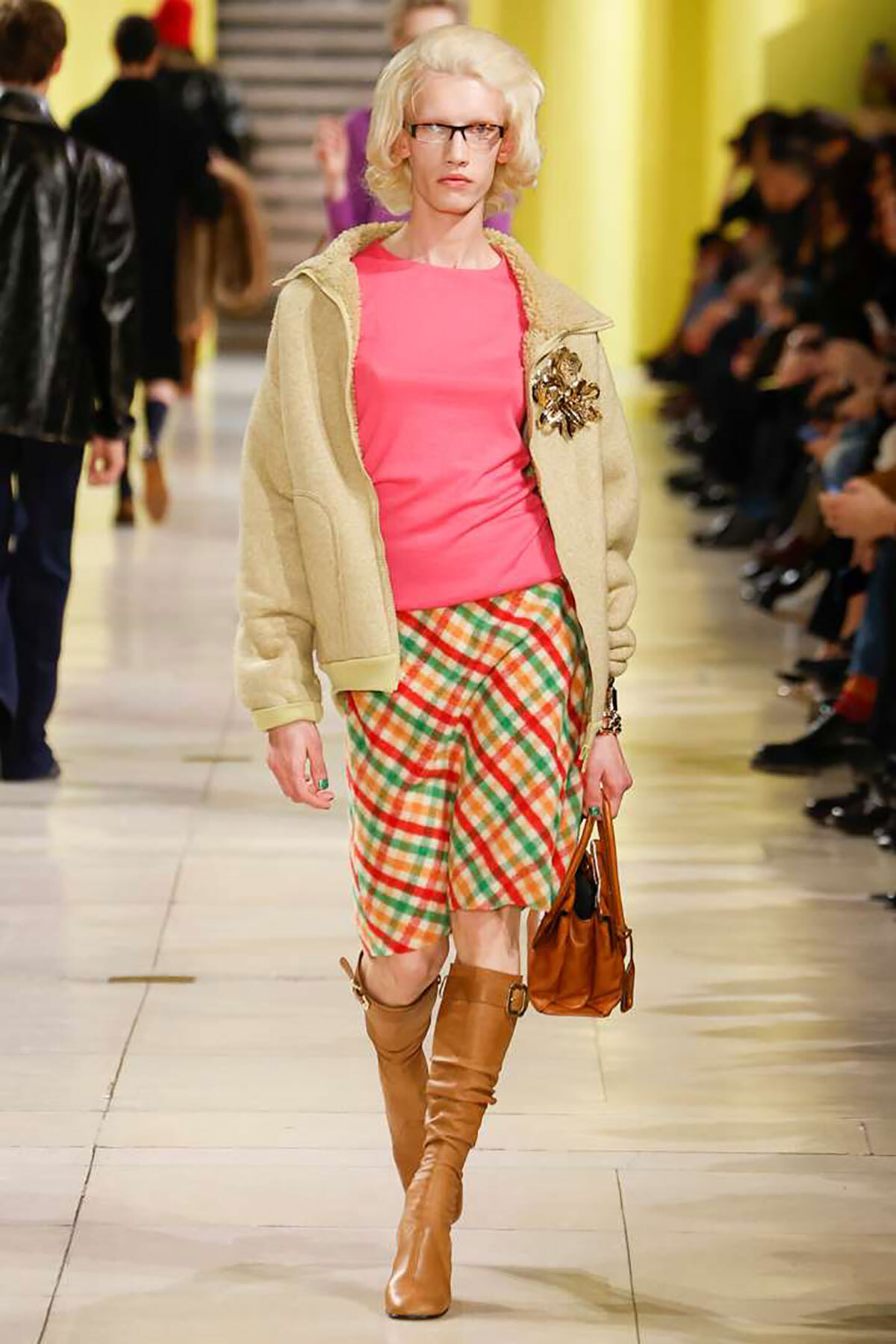 Female model on a fashion runway wearing a pink top, beige jacket, plaid shorts, tan knee-high boots, and a brown handbag.