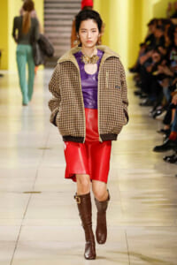 Model walks the runway in a brown plaid bomber jacket, a purple top with a circular neckline, and a bright red leather skirt, paired with knee-high brown boots.