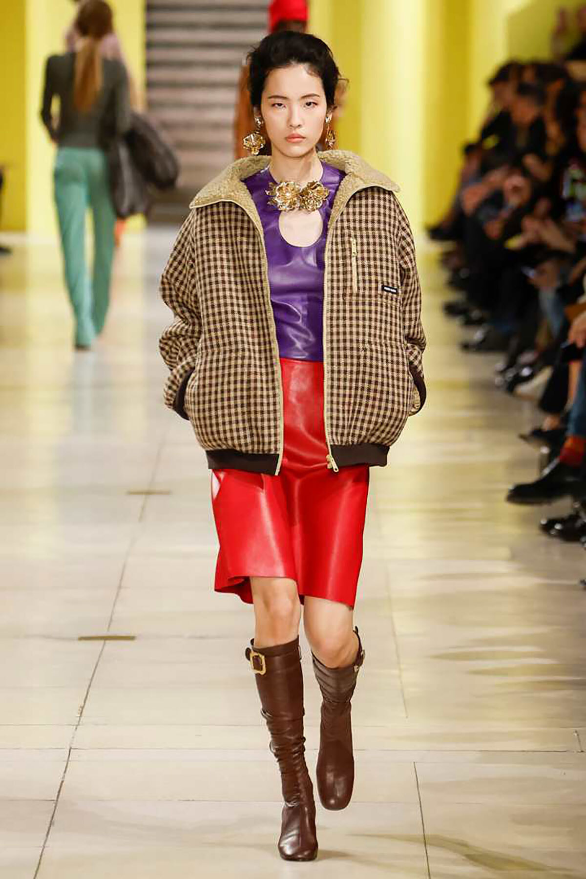 Model walks the runway in a brown plaid bomber jacket, a purple top with a circular neckline, and a bright red leather skirt, paired with knee-high brown boots.