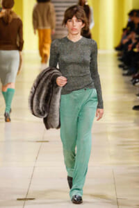 Model walking a fashion runway in a metallic long-sleeve top and mint-green trousers, carrying a fur coat over her arm.