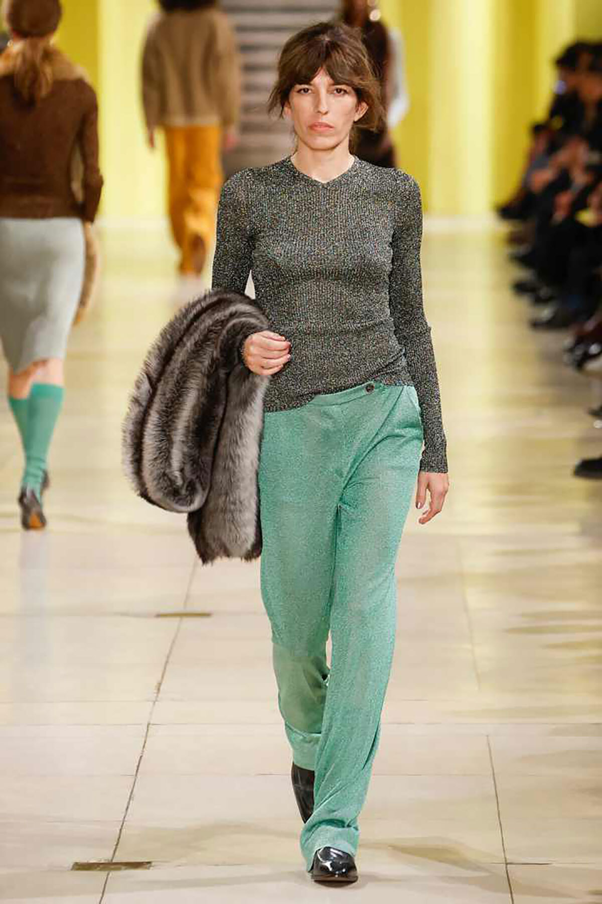 Model walking a fashion runway in a metallic long-sleeve top and mint-green trousers, carrying a fur coat over her arm.