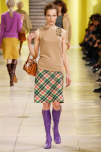 Model walking runway in beige sleeveless top, plaid knee-length skirt, and purple knee-high boots, carrying a brown handbag.
