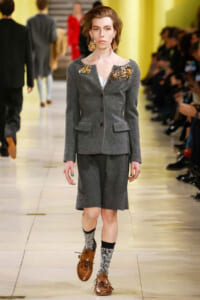 Model walks the runway in a gray tailored suit with oversized gold floral brooches on the shoulders. She wears brown lace-up shoes and sparkly socks, audience in the background.