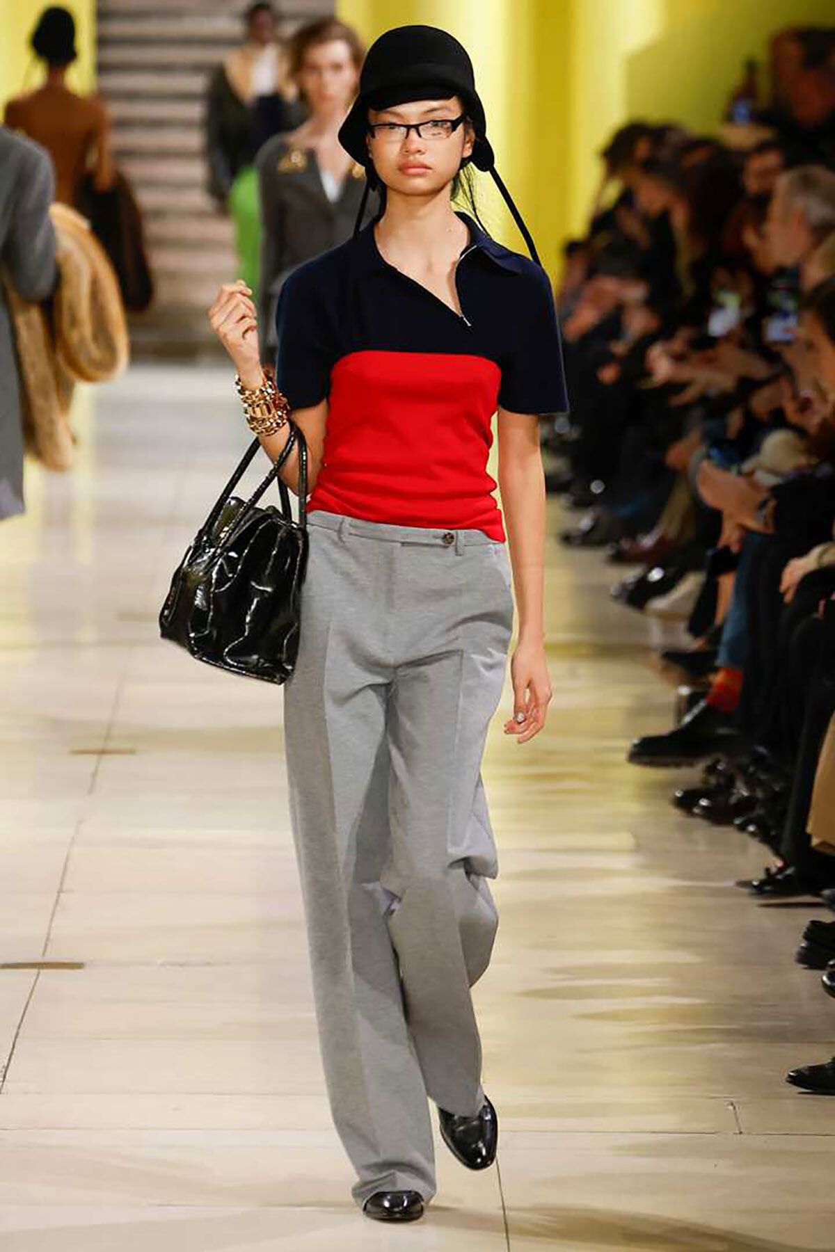 Model walking a fashion runway in a navy top with red front panel, gray wide-leg pants, black hat and glasses, carrying a black patent handbag.