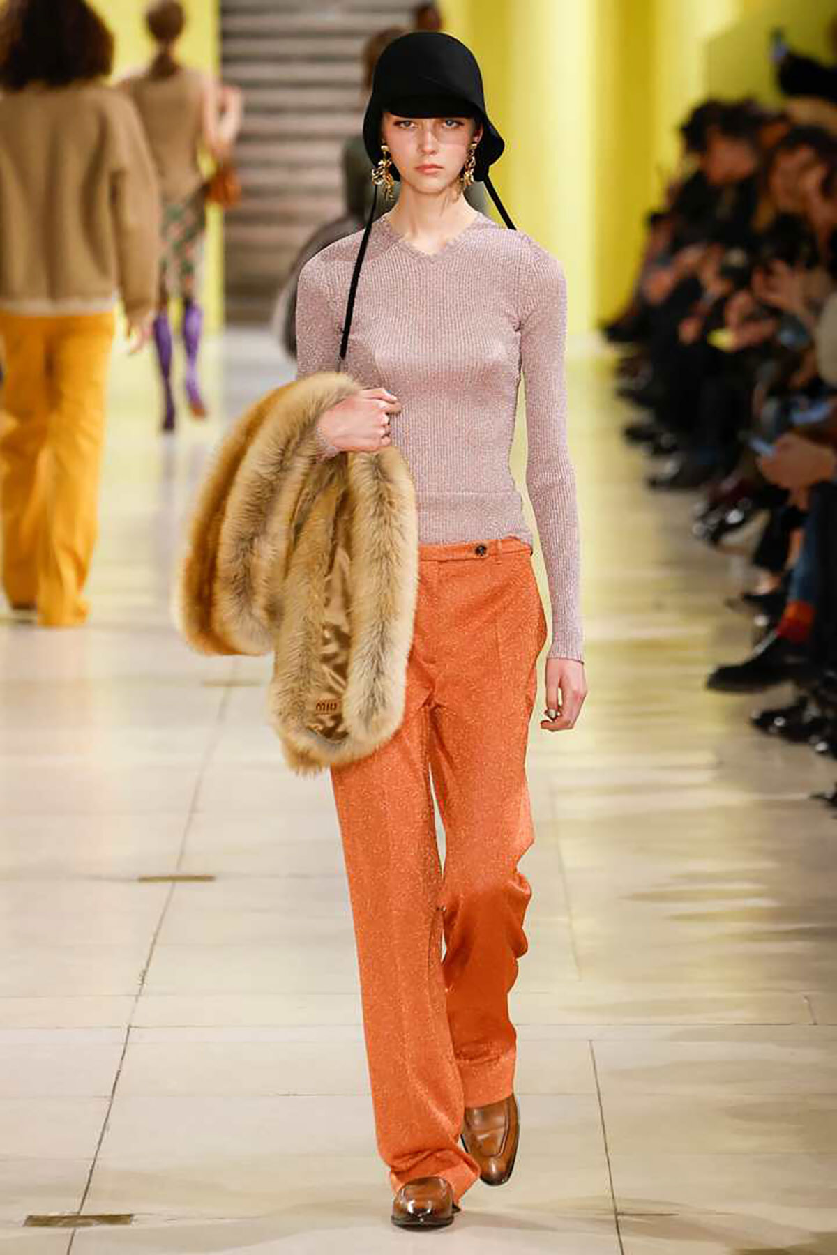 Model on runway wearing a pale pink knit top and bright orange wide-leg pants, carrying a fur coat over her arm and wearing a black bucket hat.