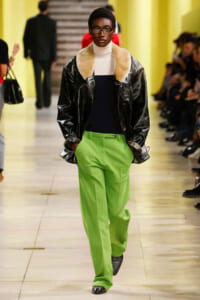 Model walks a fashion show wearing lime-green wide-leg pants, a black shiny leather jacket with a beige fur collar, and a white turtleneck.