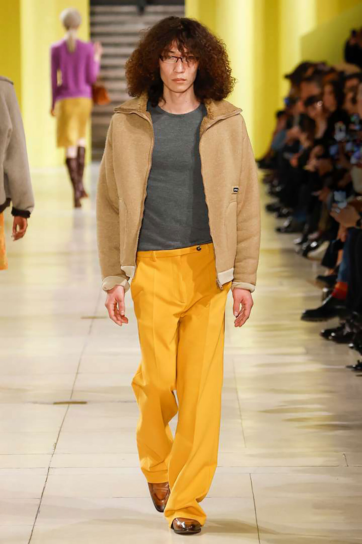 Male model walks a fashion runway in a beige zip-up jacket, gray T-shirt, and mustard-yellow pants, with audience on the right.
