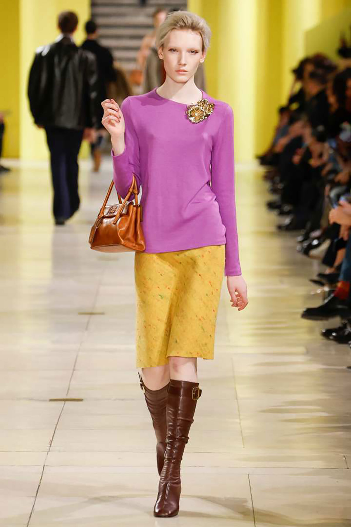 Model walking a fashion show in a purple top with a gold brooch, yellow skirt, brown knee-high boots, and a brown handbag on a bright runway.