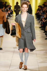 Model walks a fashion show wearing a gray wool coat over a beige top, light blue knee-high socks, brown loafers, and holding a fur stole with a black handbag.