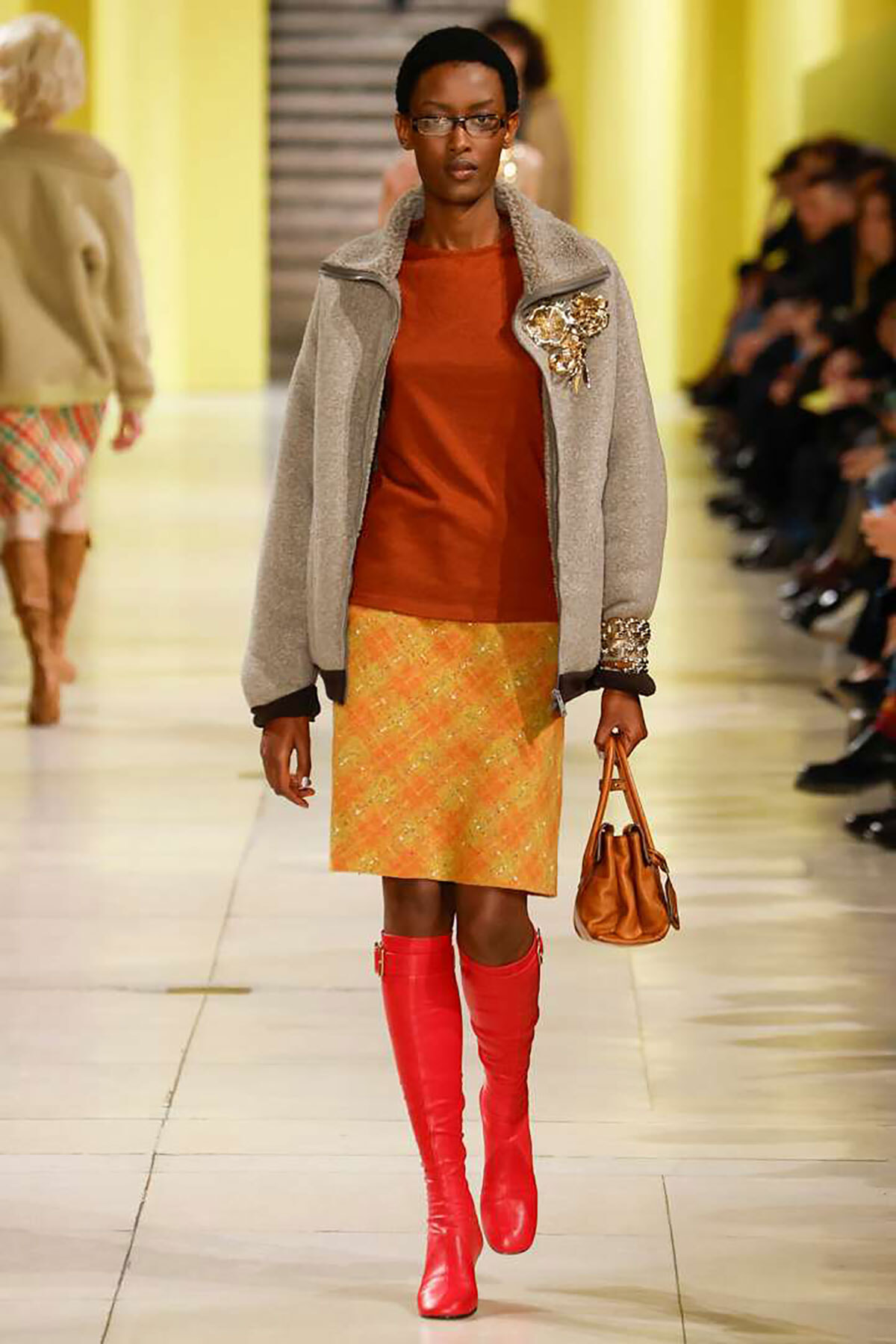 Model walking a fashion runway in a gray fleece jacket with brooch, rust-orange top, yellow-orange patterned skirt, red knee-high boots, and a brown handbag.