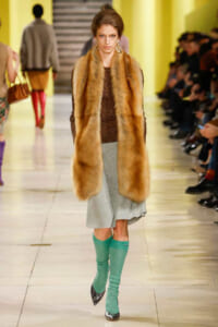 Model walking a runway in a long brown fur vest over a sequined top, silver midi skirt, and turquoise knee-high socks with pointed heels.