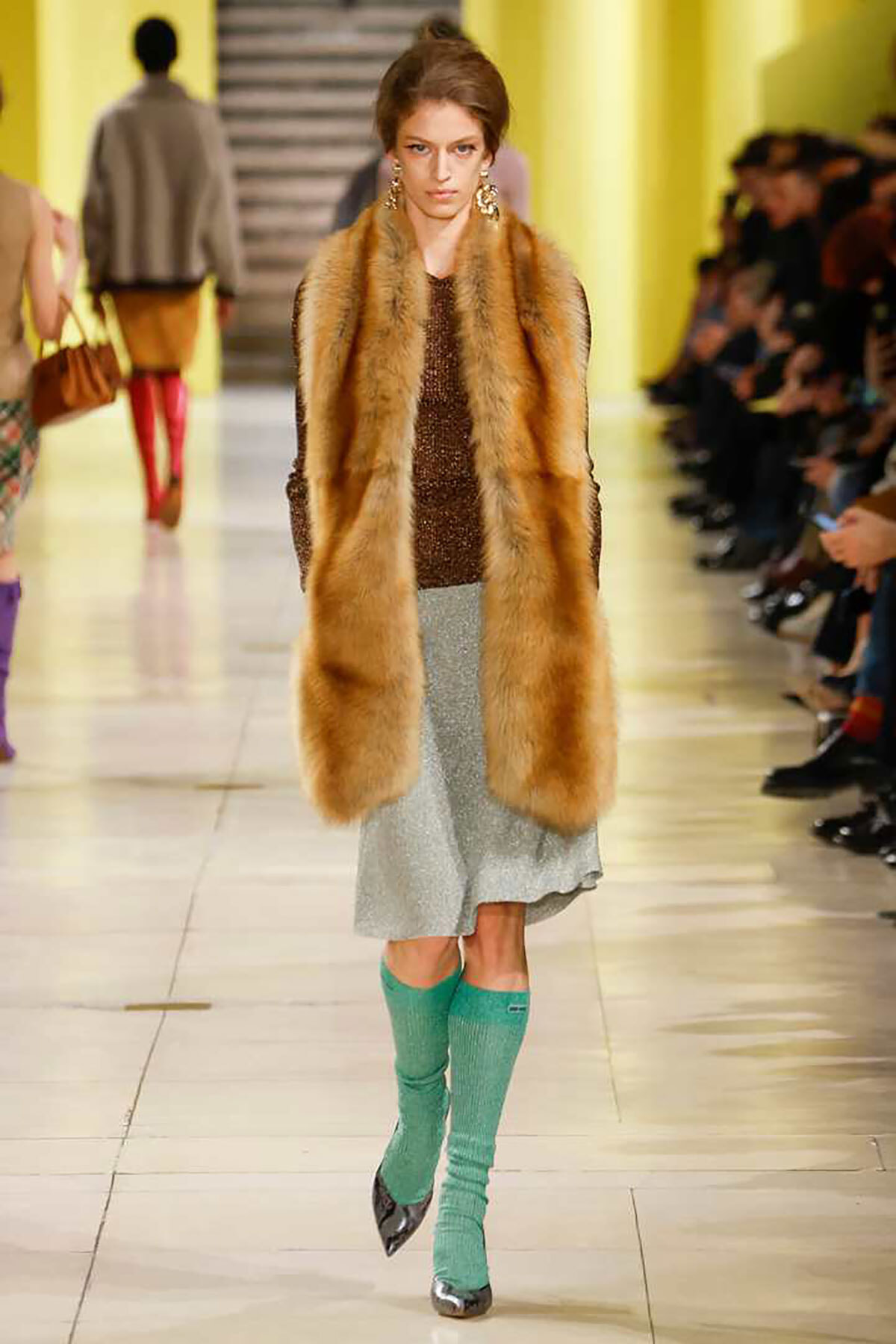 Model walking a runway in a long brown fur vest over a sequined top, silver midi skirt, and turquoise knee-high socks with pointed heels.