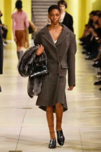 Model walking a runway in a gray double-breasted wool coat, wearing tan legwear and black loafers, carrying a fur stole and a glossy black handbag.