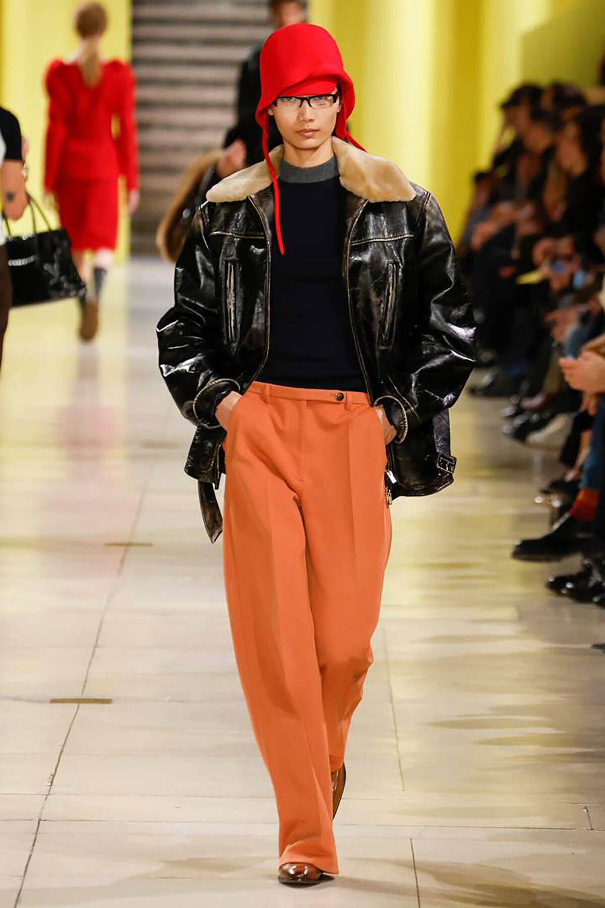 Fashion-model runway look: red hat, black leather jacket with fur collar, navy top, and wide orange pants.