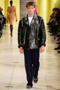 Male model on a runway wearing a shiny black leather jacket, gray turtleneck, navy pants, and silver shoes.