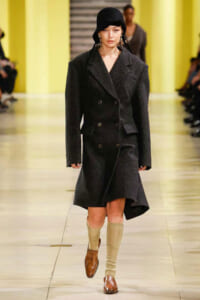 Model walking the runway in a charcoal double-breasted wool coat, black cloche hat, cream knee-high socks, and brown loafers.