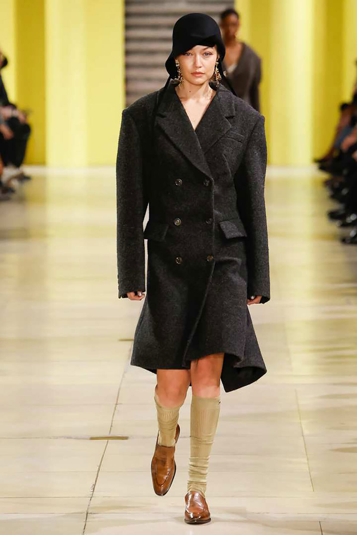 Model walking the runway in a charcoal double-breasted wool coat, black cloche hat, cream knee-high socks, and brown loafers.