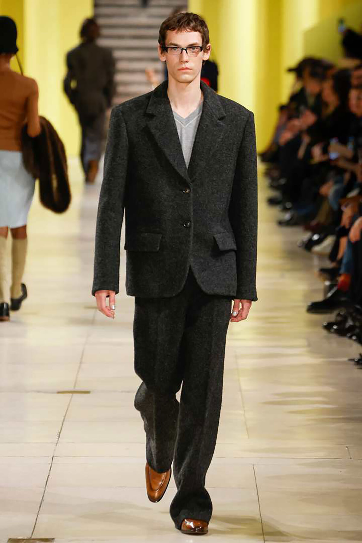 Male model on a runway wearing a dark grey textured suit with a light gray V-neck tee; brown loafers, yellow walls in the background.