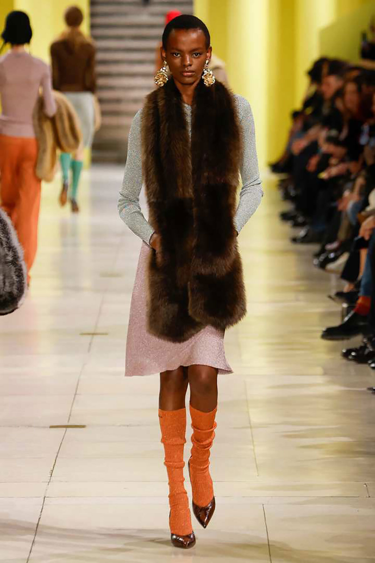 Model on a runway wearing a long brown fur vest over a silver sparkly dress, with bright orange knit leg warmers and brown pointed heels.